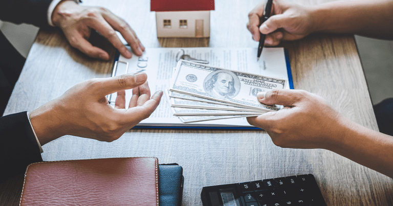 A hands a stack of cash to another person wearing a suit while they sit next to a model house.